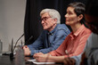 © Seventyfour - Aged Caucasian man with gray hair and glasses sitting at table with microphones, listening attentively next to middle aged Caucasian woman during talent competition judging