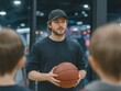 © alsu0112 - Young male basketball coach wearing a black cap is instructing children in a gym, holding a basketball while demonstrating techniques and engaging with the young players