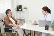 © Ekkasit A Siam - Senior asian woman in wheelchair waits calmly while orthopedic doctor reviews notes and prepares joint pain treatment plan inside clinic consultation room with books and medical items on shelf
