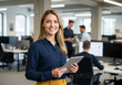 © AbdullahAl - Confident businesswoman smiling and holding a tablet in a modern office with colleagues working in the background