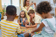 © lordn - Preschool children and teachers sitting together, threading colorful beads to create necklaces, developing fine motor skills, creativity, and hand-eye coordination in a fun classroom activity