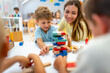 © lordn - Smiling preschool boy playing with colorful wooden blocks, guided by a cheerful teacher, in a bright and lively classroom with other children in the background