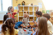 © lordn - Preschool teacher leading interactive circle time, children raising hands and smiling during an engaging early learning activity