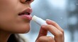 © Valiantsin - Womans hand applies white lip balm stick to dry flaky lips against blurred wintery background
