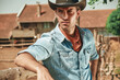 © Teodor Lazarev - A young cowboy leans against a wooden fence at a ranch, wearing a denim shirt and cowboy hat. The golden hour light enhances the warm tones of the rural landscape behind him