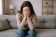 © The Little Hut - Woman Experiencing Sadness or Anxiety with Hands Covering Face, Sitting on Couch in Living Room