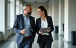 © feng - Full body vertical portrait of walking mature Latin businessman and European businesswoman discussing project on tablet in office. Two diverse partners of professional business people work together.
