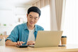 © Monster Ztudio - Happy young Asian man working on laptop in living room. Freelance worker male sitting at desk writing notes while watching webinar, studying online