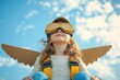 © Khusi - Joyful child wearing aviator goggles and a pilot cap with cardboard wings attached, looking upwards against a bright cloudy sky, expressing imagination and playfulness