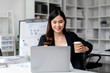 © Katcha - Asian Businesswoman Working on Laptop with Coffee Cup in Modern Office. Young Professional Enjoying Work at Desk. Business Woman Concept for Website and Marketing