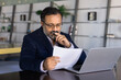 © fizkes - Thoughtful frowning senior Indian businessman reading paper document at laptop, working at computer in co-working office, reviewing invoice for tax payment, touching chin