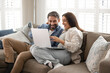 © fizkes - Couple in love relaxing on comfortable couch with laptop, enjoy online shopping through marketplace platform or websites, planning vacation, buying tickets, booking hotels, renting apartment remotely