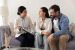 © fizkes - Friendly real-estate agent sits on sofa with couple, explains contract details, presenting information on clipboard during collaborative discussion at home. Professional services, agreement, insurance