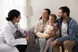 © fizkes - Friendly Indian pediatrician woman wearing white coat and stethoscope engaging in conversation with family and little 3s daughter sits on mommy lap holding favourite toy during therapist visit at home