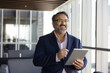 © fizkes - Cheerful happy older Indian business leader man using financial web service on tablet in office lobby, looking away, smiling, laughing, thinking on successful trading, income from investment