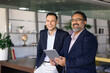 © fizkes - Happy younger and older diverse businessmen standing in office co-working space together, looking away, laughing, holding digital tablet, enjoying creative collaboration, teamwork