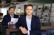 © fizkes - Positive confident young Latin vacancy applicant man posing at meeting table with older male Indian employer behind, looking at camera, getting new job in diverse company