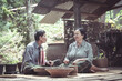 © pichet - An Elderly Couple Engaging in Traditional Conversations About Daily Life in Rural Setting Surrounded by Nature and Handmade Crafts