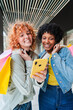 © Jose Calsina - Two cheerful young women are happily browsing their smartphone together while enjoying their shopping experience with colorful bags by their side, creating a delightful and engaging moment.