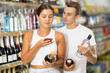© JackF - Man with wife near display of alcohol drinks in hypermarket doubt choice. Spouses buyers scan QR code, compares item in store and in mobile application, customers take pictures of drink label