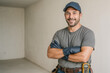 © Nargiz - Smiling male electrician wearing a navy cap, grey t-shirt, gloves, and tool belt, standing with arms crossed in a bright unfinished room, symbolizing skilled trades, electrical work