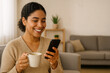 © diora - A  woman smiling at her smartphone with holds a mug. Casual home woman with her smartphone screen enjoy life.