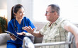 © NAMPIX - Smiling nurse in blue scrubs talking with elderly man using a walker, providing healthcare advice and support at home.
