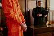 © AnnaStills - Caucasian young adult man in orange jumpsuit standing in handcuffs before judge in courtroom, judge holding folder and standing behind bench, legal proceedings in progress