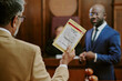 © AnnaStills - Middle aged Caucasian man holding evidence bag while standing in courtroom, Black man in suit standing in background, both participating in legal proceedings, judge visible behind