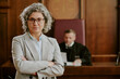 © AnnaStills - Portrait of middle aged Caucasian woman standing with arms crossed in courtroom foreground, judge sitting at bench in background, both wearing professional expressions