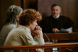 © AnnaStills - Caucasian teenager sitting at witness stand in courtroom, resting chin on clasped hands, listening attentively while judge in robe observing from bench in background