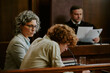 © AnnaStills - Caucasian middle aged woman sitting beside Caucasian teenage boy at courtroom table, both focusing on legal proceedings while male judge reading documents in background