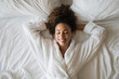 © Mediaphotos - Young adult Black woman lying on bed with eyes closed, smiling and relaxing with hands behind head, wearing bathrobe, surrounded by bedding, enjoying peaceful morning
