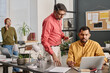 © TrueFrame Collective - Selective focus shot of Indian men wearing smart casual outfits looking at laptop screen discussing presentation design, their colleague talking on phone in background