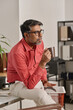 © TrueFrame Collective - Vertical medium long shot of mature Indian man drinking coffee and looking away while sitting on desk in office