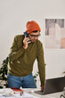 © TrueFrame Collective - Vertical medium long shot of young Indian man standing at desk in modern office talking on phone and looking at laptop screen