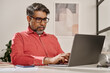 © TrueFrame Collective - Horizontal shot of mature Indian man wearing eyeglasses sitting at desk in modern office typing on laptop