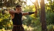 © Nabil - A muscular archer in medieval clothing, stands in the forest with sun streaming through the trees, drawing back a bow and arrow with focus and determination for hunting or target practice.
