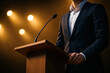 © praewpailyn - Confident man in suit delivering keynote speech at conference speaker podium with microphone under warm lights for public speaking and business presentation success
