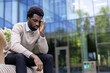 © Liubomir - A young professional man overwhelmed with stress, seated outdoors by a modern building.