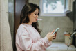 © qunica.com - A young woman multitasking, brushing her teeth with a toothbrush while checking her phone in a bathroom. The setting includes a towel, sink, and window.