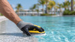 © weerasak - Person uses pool cleaning tool to maintain swimming pool, showcasing sunny day with palm trees background, evoking sense of relaxation