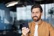 © Liubomir - A cheerful man in casual attire is using his smartphone inside a modern office. The contemporary setting includes glass walls and bright lighting, creating a professional yet comfortable atmosphere.