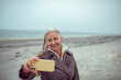 © Marko Geber - Smiling senior woman wearing coat taking a selfie on cold beach