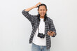 © ArvStd - A sad Asian man wearing casual clothes, backpack, camera hanging on neck, holding mobile phone, confused or frustrated expression while scratching head, isolated on white background.