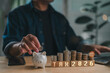 © Mdisk - Person plans future 2026 tax payments saving money. Their hand puts coin piggy bank beside growing coin stacks, symbolizing financial planning, investment, future budgeting.