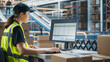 © Gorodenkoff - Caucasian Female Stocking Associate Using Desktop Computer To Check Inventory In Distribution Facility. Multiethnic Colleague Loading Cardboard Boxes With Online Orders On Automated Conveyor Belt
