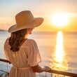 © ala - Woman in white dress and sun hat watches sunset over ocean from balcony