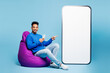 © ViDi Studio - Full body young man of African American ethnicity wears sweatshirt casual clothes sit in bag chair point on big huge blank screen area mobile cell phone smartphone isolated on plain blue background.