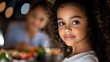 © Yuliia - Young African American girl with curly natural hair smiling warmly at dinner table with fresh healthy meal and family gathering in background, natural lighting creates cozy atmosphere.